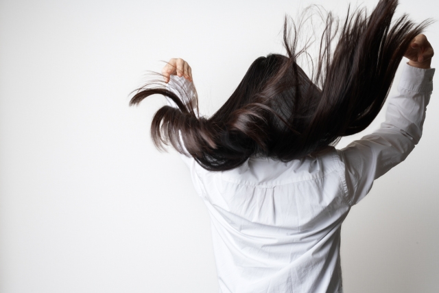 光を受けて艶やかに輝くボリュームヘアをふわりと持ち上げる女性の後ろ姿。自毛植毛のドナーヘア採取に適した健康的で滑らかな髪質をイメージしたイメージ写真
