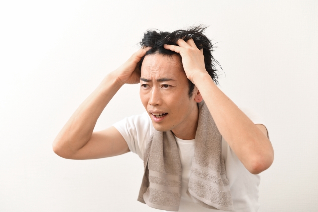 若年層の痩せ型アジア人男性が、後退し始めた生え際と髪の薄毛に悩み、頭を抱えてストレスを感じている様子。薄毛による不安や精神的ダメージ、若年男性の男性型脱毛症(AGA)対策の重要性を象徴するイメージ。