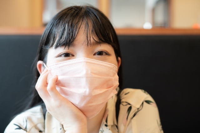 テーブルに肘をつき、顎をそっと手に預けた女性が、黒く艶やかな髪を肩から背中へと優しく流す。ピンク色のサージカルマスクが顔を半分隠していても、前髪からのぞく柔らかな目元が、優しさと穏やかさを静かに伝える。髪とまなざしが醸し出す、マスク越しの美しさと自分らしさを象徴する情景。