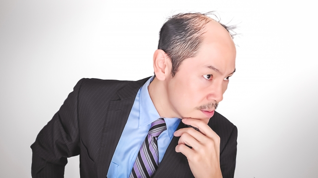 薄毛が進行し、頭皮が大きく露出している男性。フォーマルな服装で顎ひげを触れながら、スキンヘッドにするか、自毛植毛や薄毛治療薬(外用薬・内服薬)を検討するか思案している様子。男性型脱毛症(AGA)に悩む姿を表現。