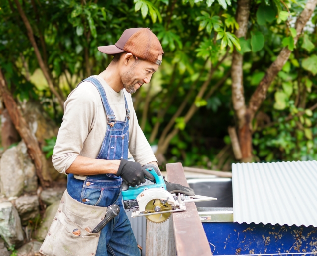 黒く整えられた髭が印象的な日本人男性が、デニムエプロンを着て楽しそうに伐採作業をしている様子。快適そうな表情が魅力的なライフスタイルイメージ