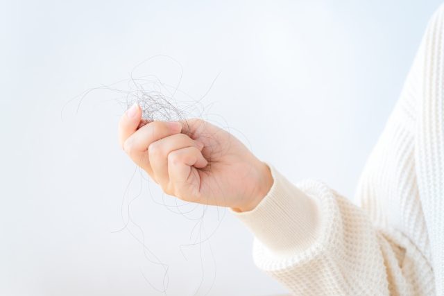柔らかなニットセーターの袖口から伸びた女性の手が、抜け落ちた細い髪の束を軽く握っている様子。抜け毛や薄毛が進行する不安や恐怖を感じさせる場面。