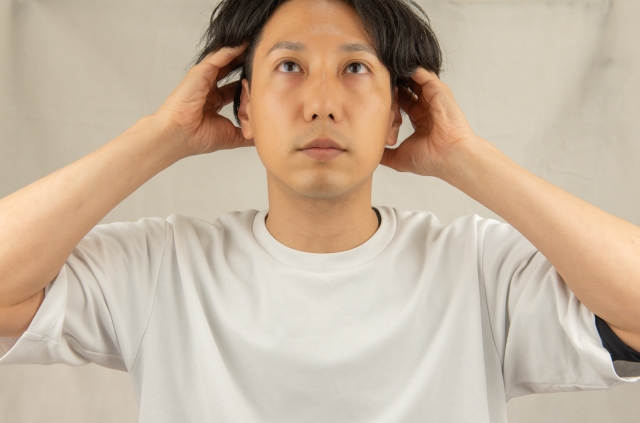 薄毛が進行し、表情も沈んだ男性が両手でこめかみ付近の髪を持ち上げている様子。頭髪のボリュームが減少し、悩みや不安を抱えている印象を与えるシーン。