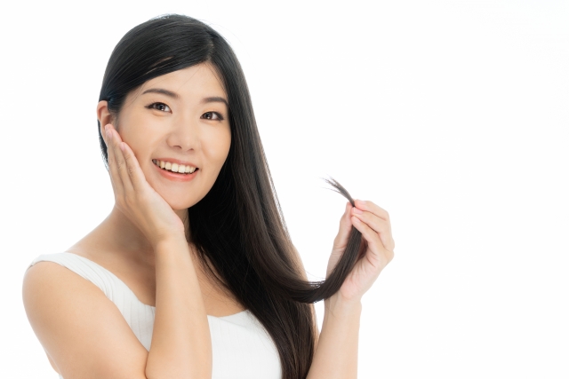 アジア人女性が髪の毛先を指先でそっとつまみながら、もう一方の手で頬を包み、優しく微笑む姿。自毛植毛におけるドナーヘアの質や状態を丁寧に確認する様子をイメージした写真