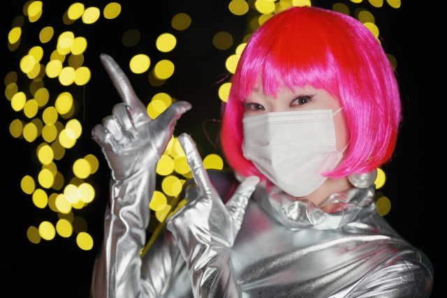 ピンクのウィッグとシルバーのボディコンドレスを身にまとい、サージカルマスクで口元を覆った女性が印象的な視線を投げかける。メイクオーバー番組での外見変化を象徴し、髪型のインパクトとイメージチェンジの重要性を示すビジュアル。