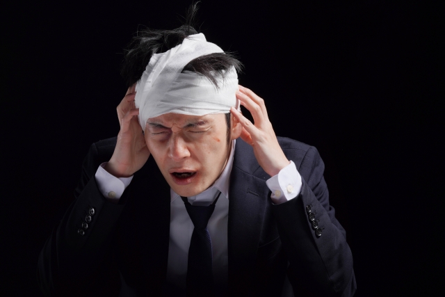 頭に白い包帯を巻き、黒髪が少し見えているスーツ姿の男性が、こめかみに手を当てて苦痛と混乱の表情を浮かべている様子