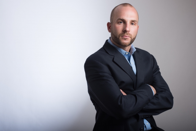 スーツ姿で腕を組み、自信に満ちた表情を浮かべる中年男性。額の生え際が後退し、頭頂部の髪が薄くなってきているが、それを堂々と受け入れている様子。口元やあご周りには濃いヒゲがあり、加齢による男性型脱毛症(AGA)の進行を自分らしく受け止めた姿。薄毛でも自信を持ち、生き方を変えない現代男性のイメージ。